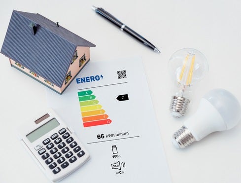 Mini model of a house next to a sheep of paper with the energy ratings on and a calculator, pen and two light bulbs on a white background