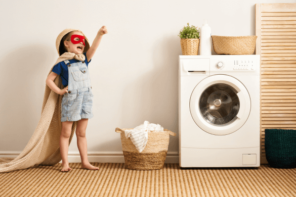 Child standing beside laundry basket and washing machine wearing hero mask and cape