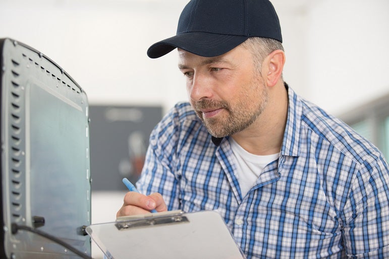 Caucasian engineer wearing a blue hat and chequered shirt writing down the appliance serial number onto his notepad