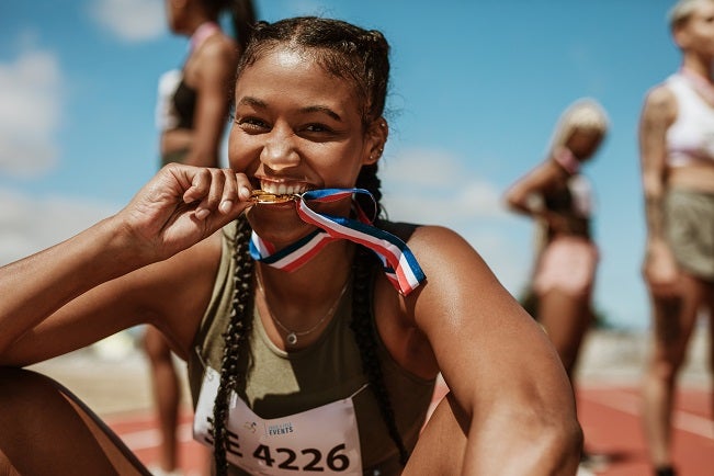 Excited mixed race female athlete biting her medal while sitting on a race track with other athletes in the background
