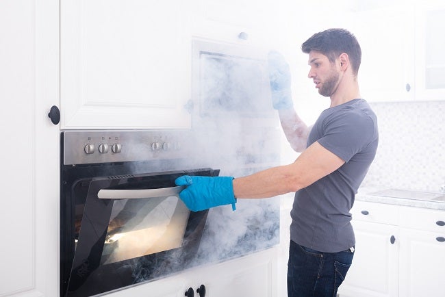 Young brunette Caucasian man wearing blue cooking gloves whilst opening the oven filled with smoke in kitchen