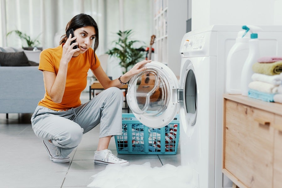 Young brunette Asian woman with a worried look on her face bent down next to a leaking washing machine whilst calling a repair service