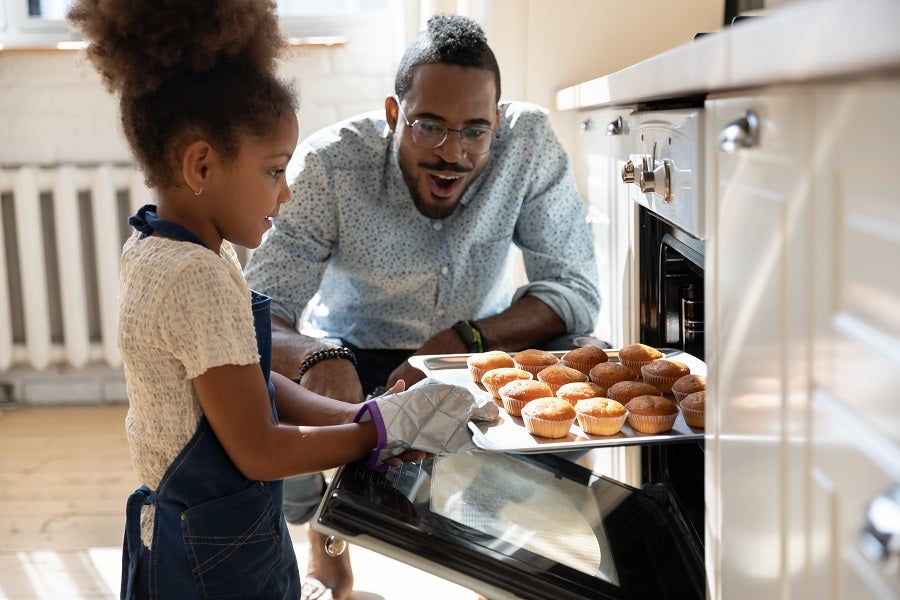 Black dad watching his young daughter taking a pan with baked tasty muffins out of the oven